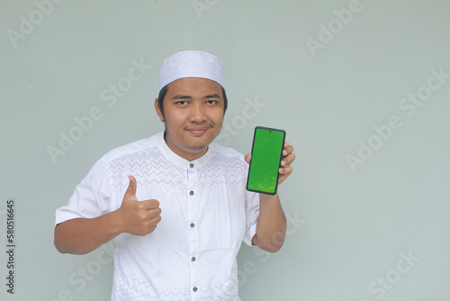 Smiling young Asian Muslim man give ok hand sign with confident gestures, while showing copy space on his phone, isolated by gray background.