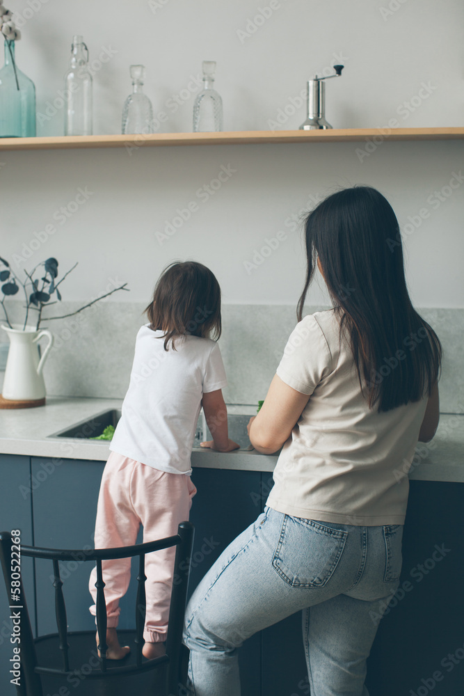 Fototapeta premium Mom and two-year-old daughter wash vegetables in the kitchen sink. Cooking and health care. Occasion moments. Back view.