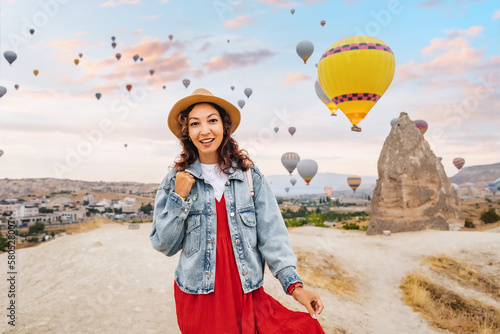 Girl traveler in her dress takes a breath of desert air, among the colorful balloons drifting through the Cappadocia sky. Basking in their beauty, she watches with a wide smile.