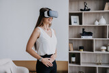 Young fit caucasian woman in white blouse and black pants using vr googles at home, playing game. Pretty Italian student girl educates by virtual reality headset. Remote education and Entertainment.