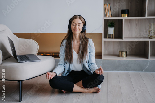 Calm confident blonde woman in blue jacket and black pants sitting on the floor legs crossed with folded hands eyes closed in meditation pose using headphones against fireplace at home. Relax at home