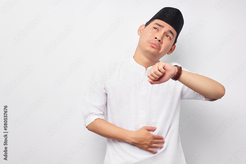 Young Asian Muslim man raised his wristwatch while touching the hand on his stomach and feeling hungry, looking aside isolated on white background. People religious Islamic lifestyle concept Young Asian Muslim man raised his wristwatch while touching the hand on his stomach and feeling hungry, looking aside isolated on white background. People religious Islamic lifestyle concept