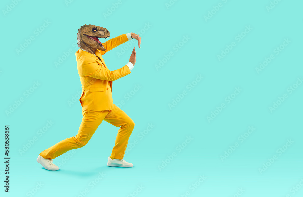 Cheerful and eccentric man in rubber dinosaur mask shows funny dance moves near copy space. Man in yellow bright suit and absurd animal mask on his head isolated on turquoise background. Full length. Cheerful and eccentric man in rubber dinosaur mask shows funny dance moves near copy space. Man in yellow bright suit and absurd animal mask on his head isolated on turquoise background. Full length.