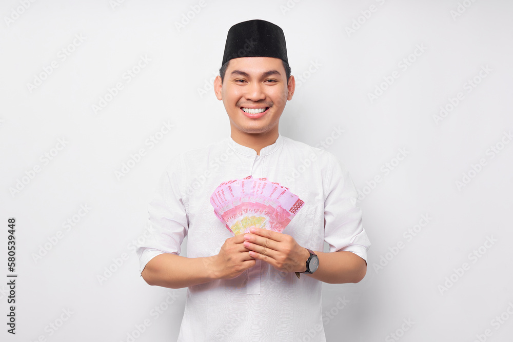 Portrait of smiling handsome young Asian Muslim man holding cash money rupiah banknotes isolated on white background. People religious Islamic lifestyle concept Portrait of smiling handsome young Asian Muslim man holding cash money rupiah banknotes isolated on white background. People religious Islamic lifestyle concept