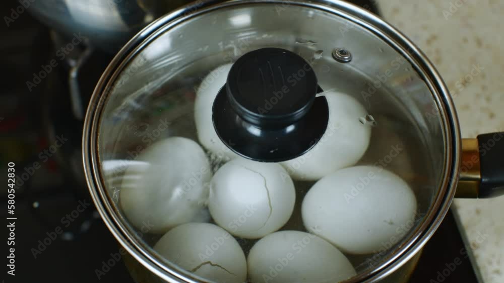 White chicken eggs are boiled in a stainless steel iron saucepan with transparent glass lid. The female hand closes a pan with boiled white chicken eggs with transparent glass roof. Close-up top view White chicken eggs are boiled in a stainless steel iron saucepan with transparent glass lid. The female hand closes a pan with boiled white chicken eggs with transparent glass roof. Close-up top view