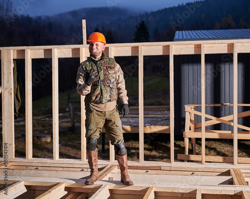 Male worker building wooden frame house. Man standing on construction site in orange safety helmet, gesturing rock and roll symbol, showing obscene horns gesture.