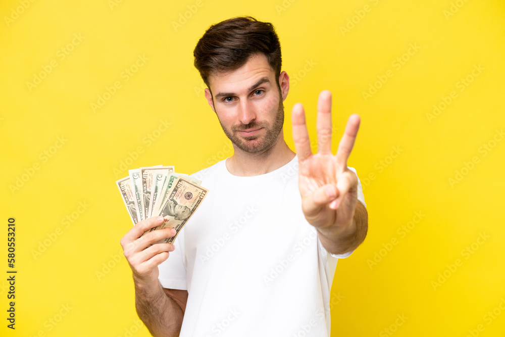 Young caucasian man taking a lot of money isolated on yellow background happy and counting three with fingers Young caucasian man taking a lot of money isolated on yellow background happy and counting three with fingers