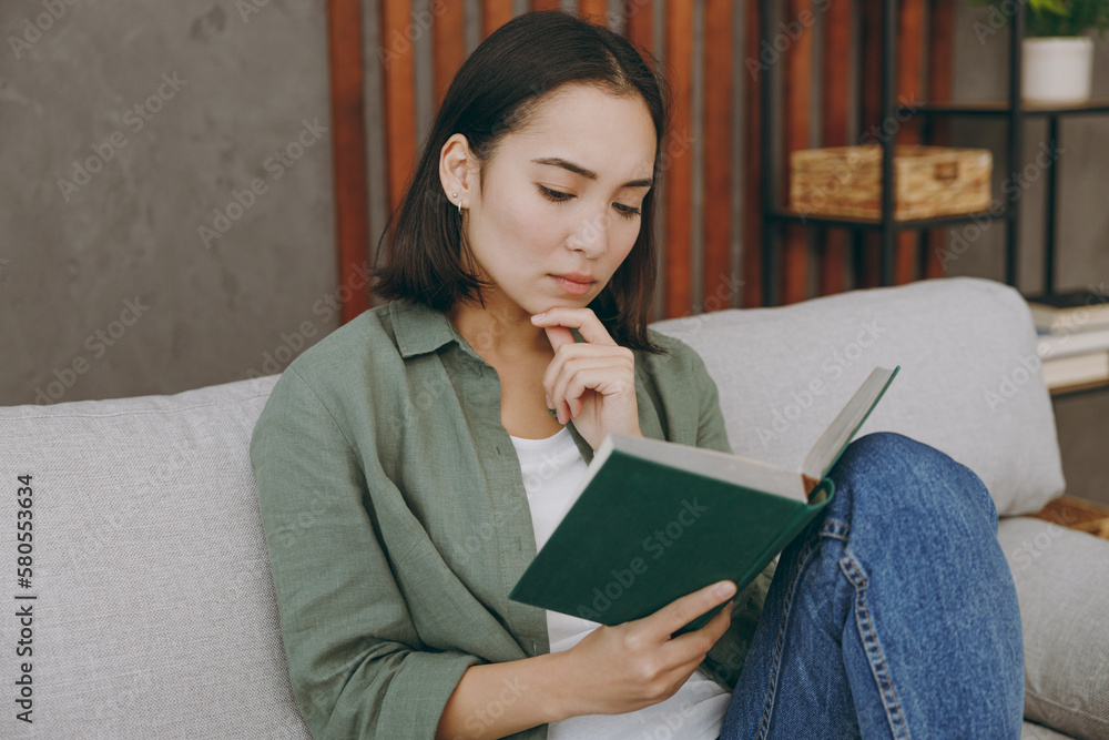 Obraz premium Young calm serious minded smart woman of Asian ethnicity wearing casual clothes read book novel sits on grey sofa couch stay at home hotel flat rest relax spend free spare time in living room indoor.