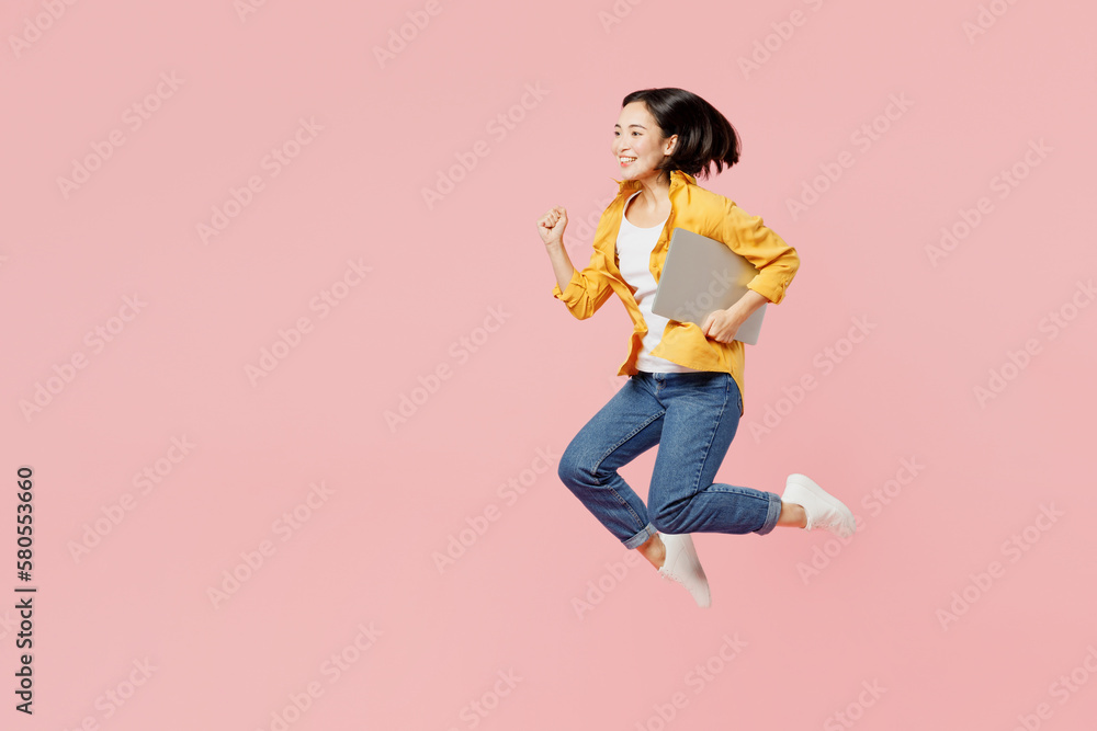Full body young satisfied cheerful cool IT woman of Asian ethnicity wear yellow shirt white t-shirt jump high hold use work on laptop pc computer isolated on plain pastel light pink background studio. Full body young satisfied cheerful cool IT woman of Asian ethnicity wear yellow shirt white t-shirt jump high hold use work on laptop pc computer isolated on plain pastel light pink background studio.