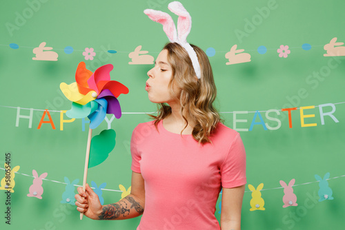 Foto Young smiling happy cheerful woman wear pink casual clothes bunny rabbit ears blowing on toy windmill isolated on plain pastel light green background studio portrait