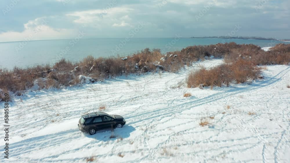 Side view Drone flying over a black car driving through a field with deep snow past bushes near Issyk-Kul Lake Side view Drone flying over a black car driving through a field with deep snow past bushes near Issyk-Kul Lake
