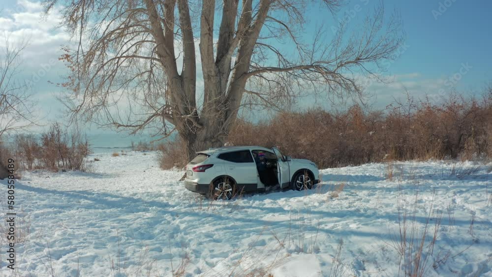 A married couple gets into a white car standing on a dirt road covered with deep snow next to a lake with a giant tree and shrubs