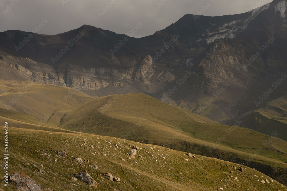 Majestic and hard summer mountain landscape - high dark gloomy mountain ridge with white glacier in overcast weather and green grass slopes in golden sunlight in highlands of Dagestan.