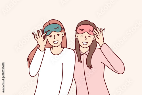 Friendly girls in pajamas with sleep masks are preparing to relax in comfortable bedroom with soft bed. Happy women use eye masks to protect themselves from sunlight during daytime naps or siestas 