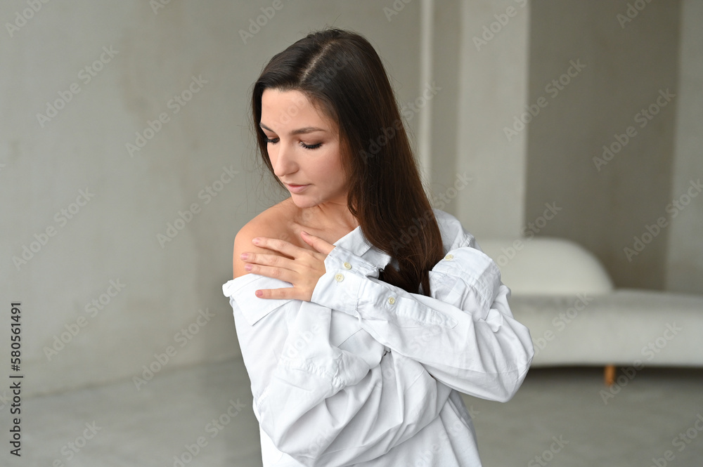 Portrait of a young dreamy brunette with closed eyes on gray background. Girl in white shirt dreaming embracing herself Portrait of a young dreamy brunette with closed eyes on gray background. Girl in white shirt dreaming embracing herself