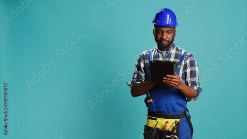 Young handyman using digital tablet on camera, preparing to start constuction contract. Male carpenter in uniform browisng online social media page on modern gadget, building site. photo