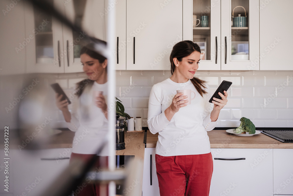Smiling woman using smart phone with coffee cup in kitchen Smiling woman using smart phone with coffee cup in kitchen