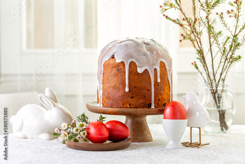 Easter cake or sweet bread decorated with white icing, red coloured Paschal eggs, spring flowers, white wooden table and window background. Easter treat, holiday decorations, symbols: bird and bunny. photo