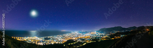 Terracina, Italy. Top View Skyline Cityscape City In Evening Night Illuminations. Panorama, Panoramic View. Amazing Bright Blue Night Starry Sky With Full Moon Above Latina Province.