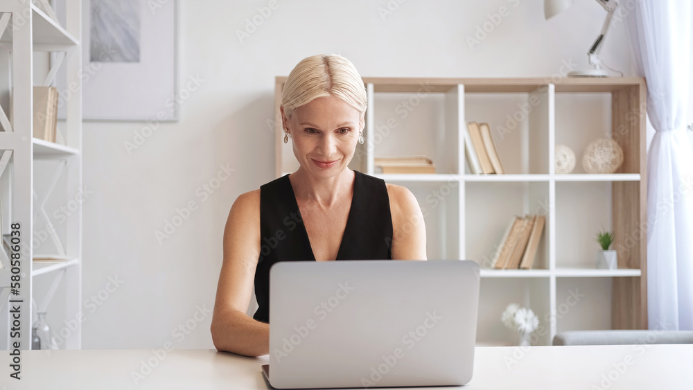 Online work. Remote job. Freelance lifestyle. Confident inspired mature business woman using laptop at light modern home office interior with free space. Online work. Remote job. Freelance lifestyle. Confident inspired mature business woman using laptop at light modern home office interior with free space.