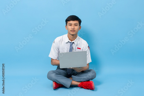 Student of Indonesian high school sitting on the floor using laptop isolated on blue background