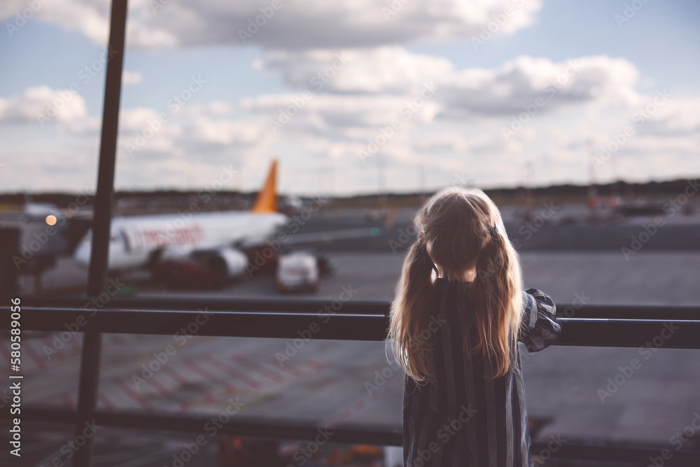 Little girl at the airport waiting for boarding at the big window. Cute kid stands at the window against the backdrop of airplanes. Looking forward to leaving for a family summer vacation