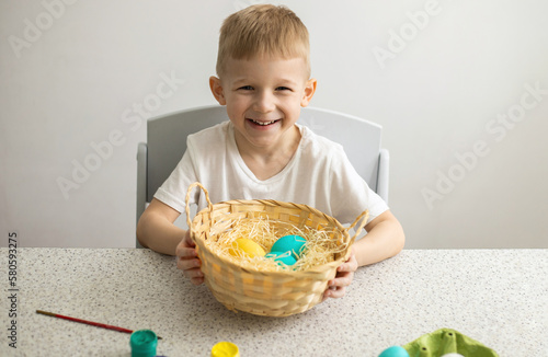 A happy child sitting at a table on a white background holds a wicker basket with painted eggs. There are brushes and paints on the table. Easter.