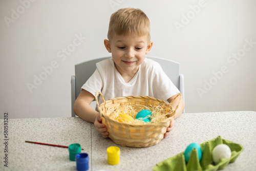 A child sitting at a table on a white background holds a wicker basket with painted eggs. There are brushes and paints on the table. Easter.