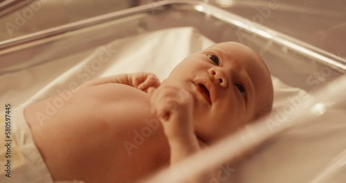 Cute Little Caucasian Newborn Baby Lying in Bassinet in a Maternity Hospital. Close Up Face Portrait of a Tiny Playful and Energetic Child. Healthcare, Pregnancy and Motherhood Concept. Warm Color photo