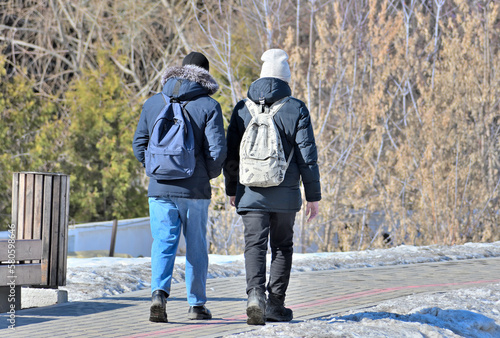 Two men with backpacks are walking along a park alley on a spring day