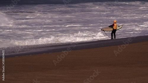 Surfer in yellow vest and helmet with board, ready to go in the water , standing on the shore in front of wild waves
