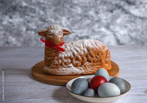 A cupcake in the form of the lamb of God for Easter with a plate of painted eggs against a gray marble wall. Festive treats