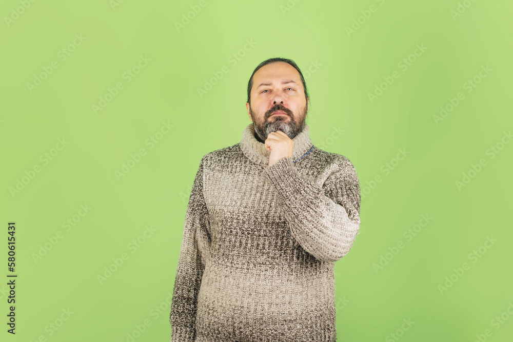 A serious middle-aged man with a beard in a warm sweater holds his chin thoughtfully. Warmly dressed man on a plain background. Background on the theme of deep reflection. A serious middle-aged man with a beard in a warm sweater holds his chin thoughtfully. Warmly dressed man on a plain background. Background on the theme of deep reflection.