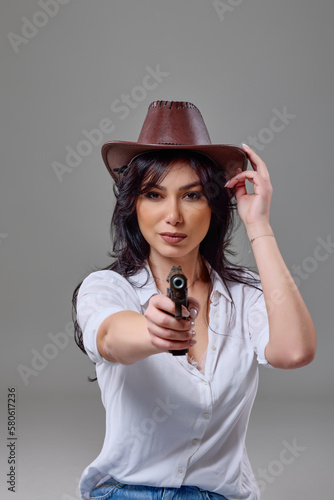 a beautiful woman with a cowboy hat and a gun in her hand posing in the studio on a gray background