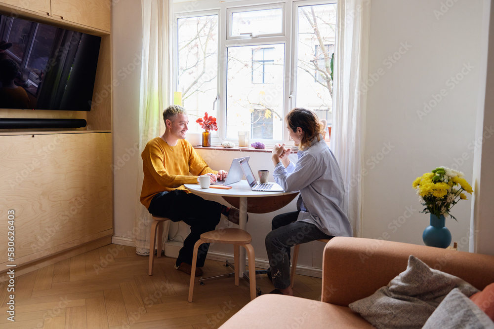 Same Sex Male Couple In Lounge Sitting At Table With Laptop Working From Home Same Sex Male Couple In Lounge Sitting At Table With Laptop Working From Home
