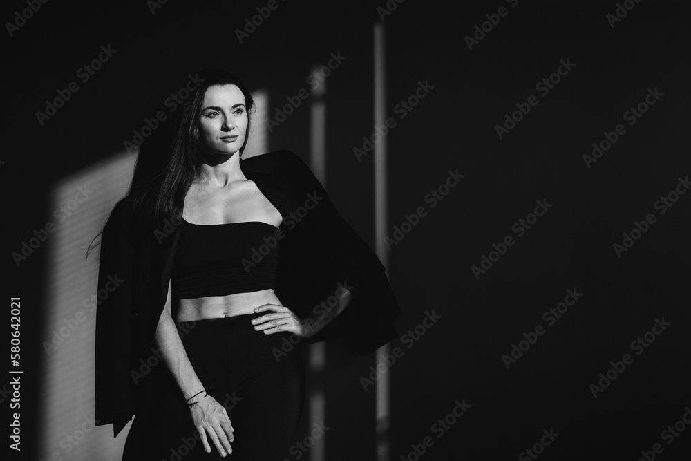 A beautiful brunette poses against the background of a gray wall in a black jacket and top, light from the window. BW photo.