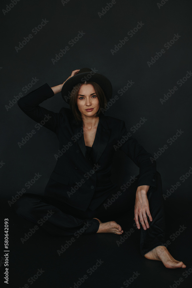 Portrait of a modern stylish woman. A brunette in a suit and hat poses barefoot on a black background. Portrait of a modern stylish woman. A brunette in a suit and hat poses barefoot on a black background.