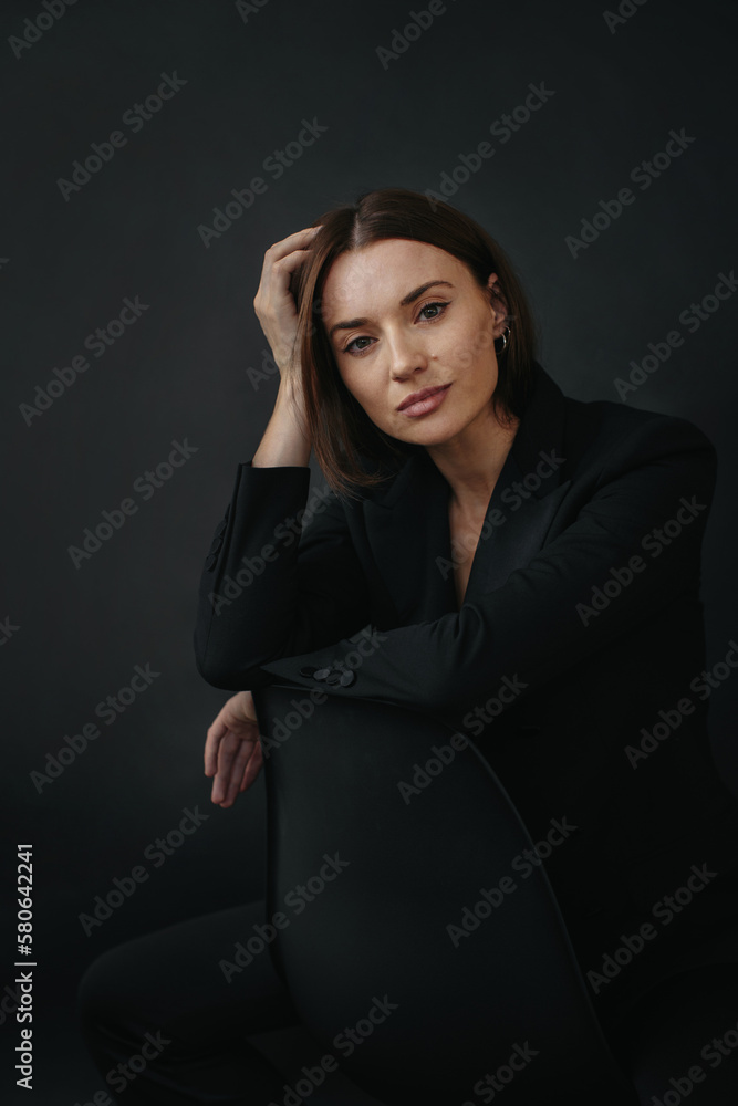Naklejka premium Portrait of a modern stylish woman. Beautiful brunette woman posing on a chair on a black background.