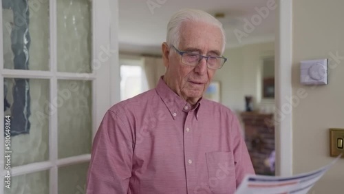 Worried Senior Man With Bill Turning Down Central Heating Thermostat At Home In Energy Crisis