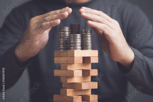 Risks in business or financial concept. Idea to prevent risk in business. Business man using hand to protection coin stacked on tower wooden block game and prevent falling down. © SKT Studio