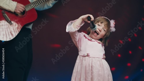 Little girl on stage in vintage dress, she sings into microphone and dances, her father plays acoustic guitar. Color music is shining. Performance on stage. Early development of children