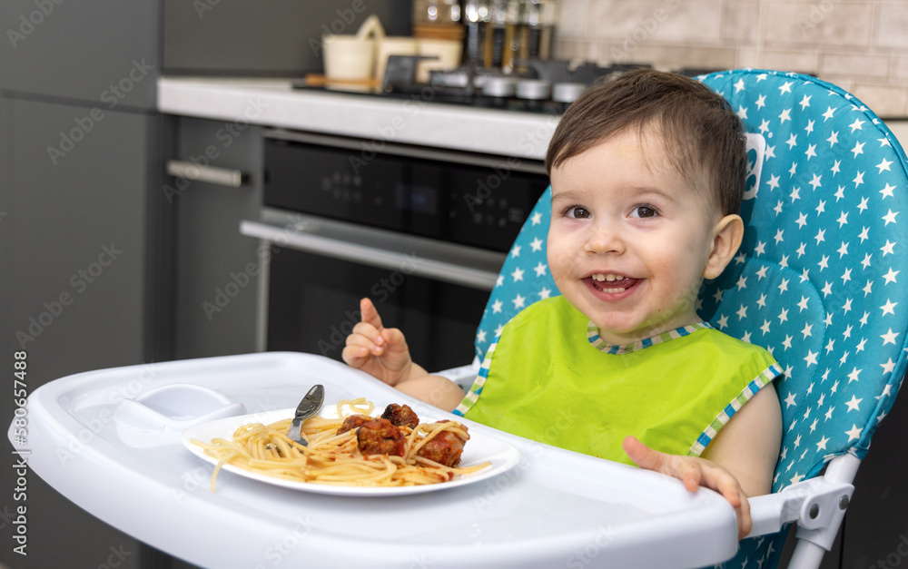 child baby boy eating pasta spaghetti with meatballs and tomatoes sauce sitting in high chair kitchen background.kid toddler putting food with both hand in mouth hungry cute infant.diversification