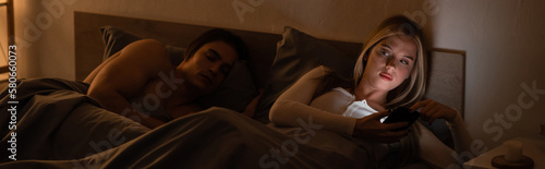 Cuadro en lienzo blonde woman messaging on smartphone next to sleeping boyfriend at night, banner