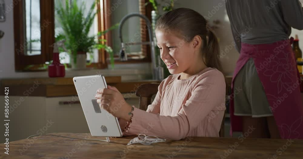 Portrait of a Cute Little Girl Using a Digital Tablet Computer at Home in the Kitchen, While Mother is Cooking in the Background. Female Kid Playing Educational Games and Using Technology Portrait of a Cute Little Girl Using a Digital Tablet Computer at Home in the Kitchen, While Mother is Cooking in the Background. Female Kid Playing Educational Games and Using Technology
