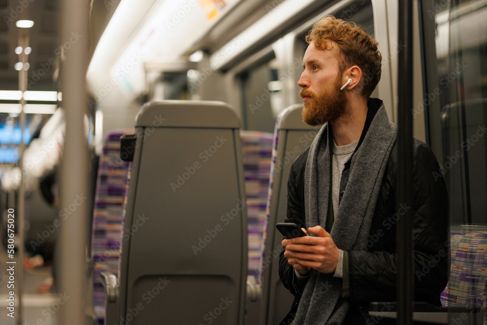Man is sitting in a subway train with a smartphone in his hands and wearing wireless headphones Man is sitting in a subway train with a smartphone in his hands and wearing wireless headphones