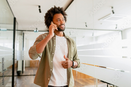 Male office employee having phone conversation, connecting with customers. Confident African-American man in smart casual wear talking on the smartphone while walkiing at the coworking office photo