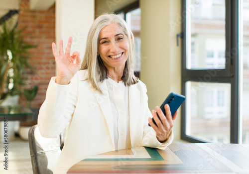 pretty senior woman feeling happy, showing approval with okay gesture with a smartphone