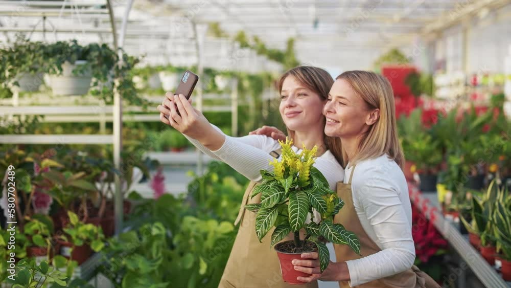 Side view of two positive young florists enjoying work in greenhouse. Women wearing uniform, taking selfie, smiling, holding flowerpot. Concept of happiness and sincere emotions.