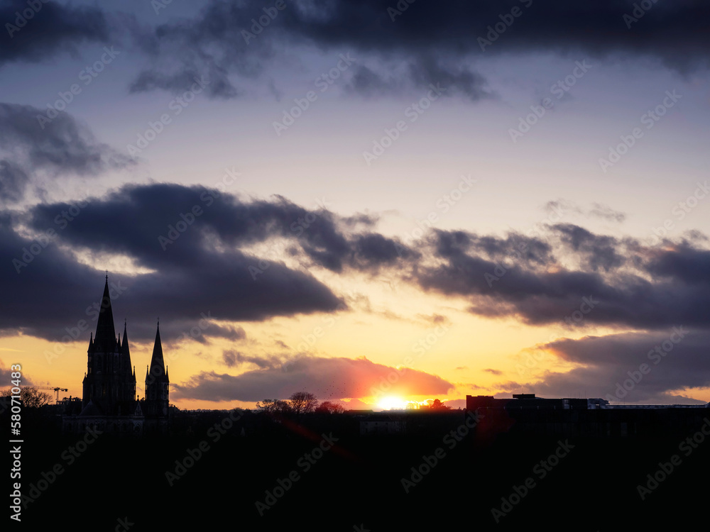Silhouette of a spire of a catholic church against dark cloudy sunset sky and setting sun
