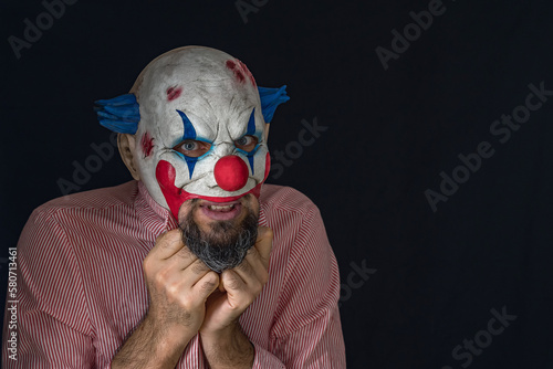 A creepy clown looks into the camera while standing in a dark room. A man wearing a scary clown mask and a red striped shirt holds onto his collar. A bearded clown maniac. © VeNN